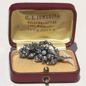 May include: A silver-toned floral brooch with sparkling clear stones, displayed in a red jewelry box with gold-colored lining. The box has the text "C.J. Jonssons" on the inside lid. The brooch features two flower clusters and delicate leaf details.