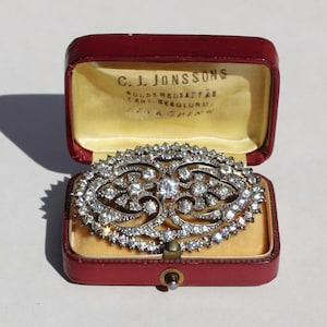 Puede incluir: Broche plateado adornado con numerosas piedras preciosas transparentes, exhibido en un joyero rojo con interior amarillo. El broche presenta un diseño simétrico con patrones arremolinados y un punto focal central.