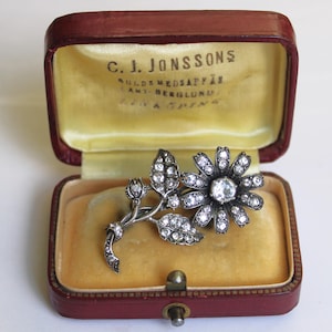 May include: A silver-toned floral brooch with clear crystals, displayed in a vintage red jewelry box with yellow velvet lining. The brooch features a large flower and leaves.