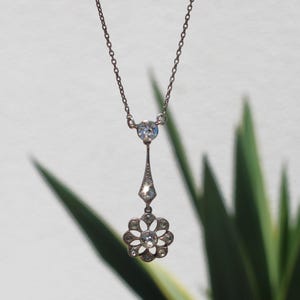 Op de afbeelding: Een delicate zilveren ketting met een bloemvormige hanger met heldere edelstenen. De hanger hangt aan een fijne ketting en bevat een ronde edelsteen aan de bovenkant. Het bloemmotief heeft meerdere bloemblaadjes, elk met een edelsteen.