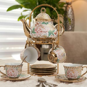 May include: A gold-colored tea set with a teapot, cups, saucers, and spoons. The teapot and cups feature a floral design with pink and green accents. The set is displayed on a gold metal stand, creating an elegant presentation.