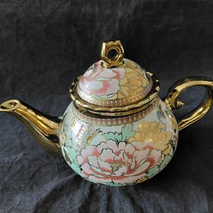 May include: A porcelain teapot featuring a floral pattern with pink, yellow, and mint green blossoms on a white background. The teapot has a gold-colored spout, handle, and lid finial, with a scalloped edge on the lid and a butterfly motif.