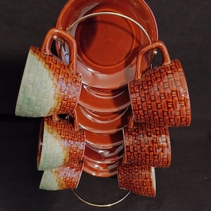 May include: A set of six brown ceramic teacups and saucers with a metal rack. The cups have a basketweave design with a two-tone color scheme of brown and light green. The saucers and the large top saucer are a solid brown color.