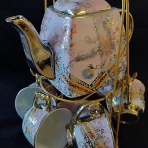 May include: A porcelain tea set featuring a teapot and matching cups with a pink base, intricate floral patterns in gold, blue, and orange, and metallic gold accents on the handles, spouts, and rims, all suspended on a gold metal wire stand.