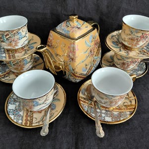 May include: A tea set featuring a square teapot and four matching cups and saucers. The ceramic pieces are decorated with a floral pattern in shades of orange, blue, and gold, with gold-toned rims and handles. Four silver-toned spoons are included.