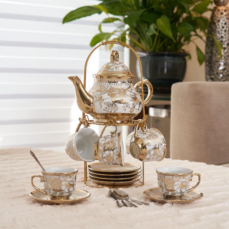 May include: A white and gold tea set with floral designs. The set includes a teapot, cups, saucers, and spoons, all displayed on a gold-colored metal stand. The teapot and cups have gold accents.