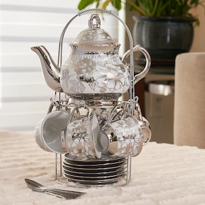 May include: A silver-plated tea set with a floral design. The set includes a teapot, six cups, saucers, and spoons, all displayed on a metal stand. The teapot and cups have a white background with silver floral patterns.