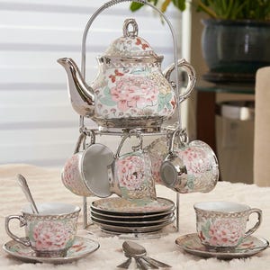 May include: A silver-toned tea set with a floral design. The set includes a teapot, six cups, saucers, and spoons, all displayed on a tiered metal stand. The cups and teapot feature a pink peony pattern with silver trim, creating an elegant aesthetic.