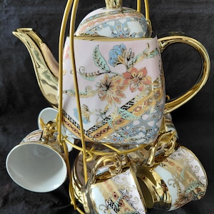 May include: A porcelain teapot and matching teacups set featuring a light pink base with intricate floral patterns in blue, orange, and green, accented by gold trim on the handles, spouts, and rims, all held within a gold-toned metal wire stand.
