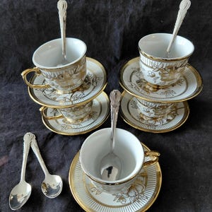May include: A set of five white porcelain teacups and saucers featuring gold floral patterns and gold-trimmed edges. Each cup includes a matching silver-toned metal spoon with a decorative handle, arranged in a stacked and individual display.