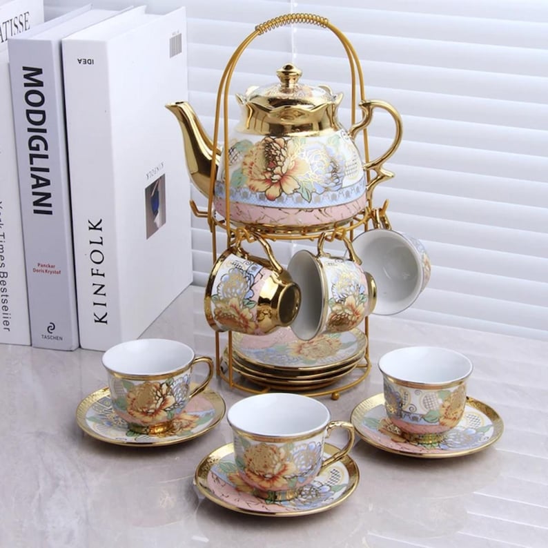 May include: A gold-colored tea set with a teapot, cups, and saucers. The teapot and cups feature floral designs with gold trim. The set is displayed on a gold metal stand, creating an elegant presentation. The cups are white inside.