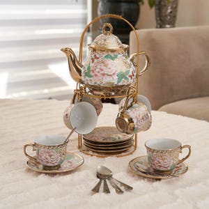 May include: A gold-colored tea set with a teapot, cups, saucers, and spoons. The teapot and cups feature a floral design with pink and green accents. The set is displayed on a gold-colored metal stand.