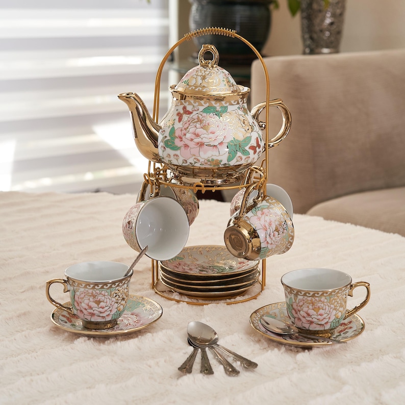 May include: A gold-colored tea set with a teapot, cups, saucers, and spoons. The teapot and cups feature a floral design with pink and green accents. The set is displayed on a gold-colored metal stand.