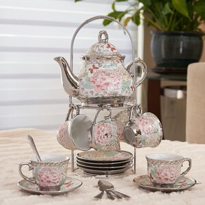 May include: A floral porcelain tea set with a silver-tone metal stand. The set includes a teapot, six teacups, saucers, and spoons. The cups and teapot feature a pink peony design with silver trim. The stand has a curved handle.