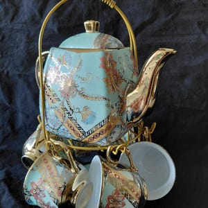 May include: A light blue teapot and matching teacups set featuring gold floral patterns and gold-toned trim. The set is arranged on a gold metal wire rack with a curved handle, showcasing a hexagonal teapot body and cups with gold handles.