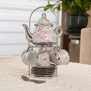 May include: A porcelain tea set featuring a teapot, four cups, and four saucers with a floral pattern of pink peonies and butterflies. The set includes a silver-toned metal stand with a handle and four matching silver-toned metal spoons.