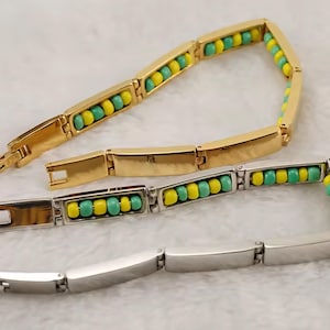 May include: Two link bracelets, one gold-toned and one silver-toned, each featuring a series of rectangular metal segments. The inner side of each bracelet displays alternating small yellow and turquoise beads set within the metal frame.