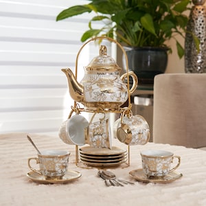 May include: A white porcelain tea set featuring gold floral patterns, including a teapot with a lid, several teacups, and saucers. The set is displayed on a gold metal stand, with additional teacups, saucers, and small silver spoons arranged below.
