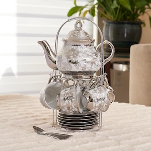 May include: A silver-toned tea set with a floral design, including a teapot, cups, saucers, and spoons. The set is displayed on a metal stand. The teapot has a lid and a curved spout. The cups and saucers are white with silver floral patterns.