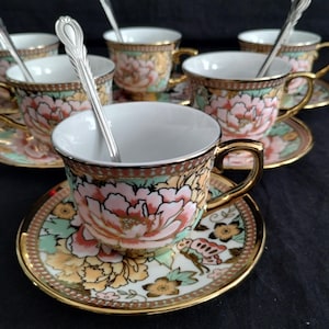 May include: A set of six floral teacups and saucers with matching spoons. The cups and saucers feature a pink peony design with gold trim. The spoons are silver. The teacups are on a black surface.