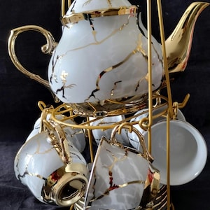 May include: A white porcelain tea set with gold accents, including a teapot, cups, saucers, and spoons. The set features a gold-colored metal stand. The teapot and cups have a gold vein design.