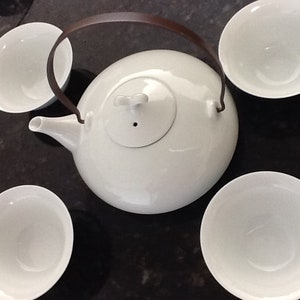 May include: A white ceramic teapot with a brown handle and four matching teacups. The teapot has a rounded shape and a spout. The teacups are small and round. The set is arranged on a dark surface.