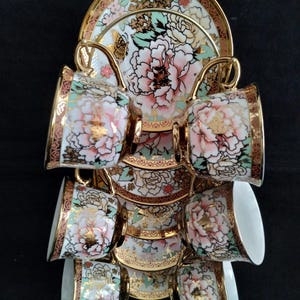 May include: A set of six teacups and saucers with a floral peony design in pink, gold, and green. The cups and saucers are displayed on a gold-colored metal rack. The teacups have gold rims and handles.
