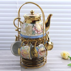 May include: A gold-colored tea set with a teapot, cups, and saucers. The teapot and cups are decorated with floral patterns in pink, white, and gold. The set is held in a gold metal rack.
