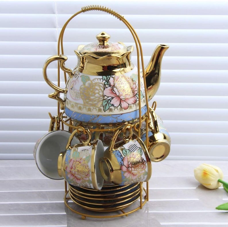 May include: A gold-colored tea set with a teapot, cups, and saucers. The teapot and cups are decorated with floral patterns in pink, white, and gold. The set is held in a gold metal rack.