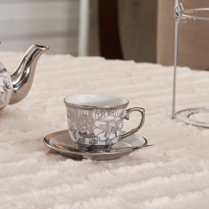 May include: A silver-plated teacup and saucer set with floral designs. The cup and saucer have a white interior and a silver exterior with intricate floral patterns. A small spoon rests on the saucer. The set is on a soft, light-colored surface.