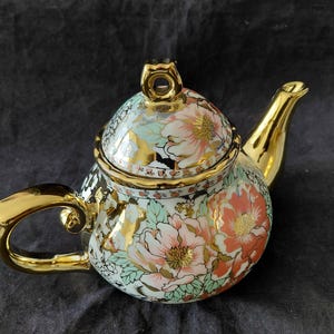 May include: A porcelain teapot featuring a white base with a floral pattern of pink and orange peonies, light green leaves, and black accents. The teapot includes a shiny gold handle, a gold spout, a gold-trimmed lid, and a gold finial knob.
