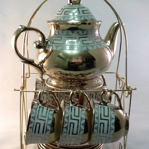 May include: A gold-colored tea set with a teapot, cups, and saucers. The teapot and cups have a white and gold geometric pattern. The set is displayed on a gold metal rack.