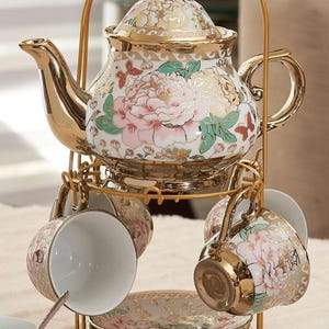 May include: A porcelain tea set featuring a teapot, cups, and saucers with a floral pattern of pink peonies and butterflies, accented with gold trim. The set is displayed on a gold-colored metal stand with a handle, holding the teapot and cups.