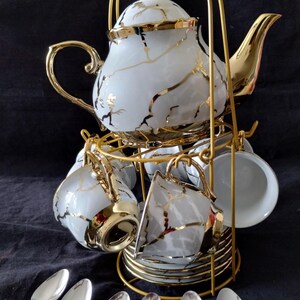 May include: A white porcelain tea set with gold accents, including a teapot, cups, saucers, and spoons. The set is displayed on a gold metal stand. The teapot and cups have a gold vein design.