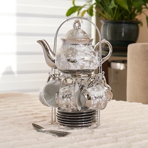 May include: A silver-toned tea set with a floral design. The set includes a teapot, cups, saucers, and spoons, all displayed on a metal rack. The teapot and cups have a white background with silver floral patterns.