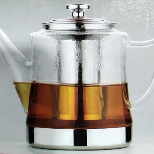 May include: A clear glass teapot with a stainless steel base, lid, and infuser. The teapot is filled with brewed tea, showing a golden-brown color. The handle and spout are also made of clear glass.