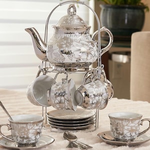 May include: A silver and white floral tea set, including a teapot, cups, saucers, and spoons, displayed on a tiered metal stand. The teapot and cups feature a detailed floral design. The set is designed for serving tea.