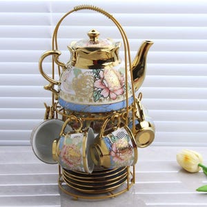 May include: A gold-colored tea set with a teapot, cups, and saucers. The teapot and cups feature floral designs in pink, white, and gold. The set is held in a gold metal rack.
