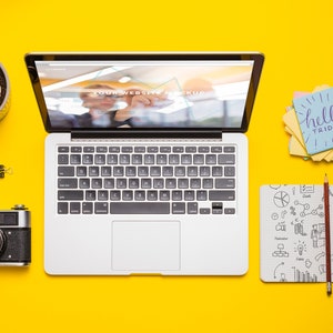 May include: A yellow desk with a laptop, a camera, a notebook, a pencil, and a stack of sticky notes with the text "hello friday".