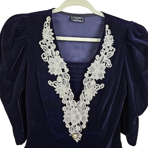 May include: A vintage navy blue velvet top with a white lace floral design around the neckline. The top features puffed sleeves and a pearl-like button. The label reads "Cachet, Kari Welles."