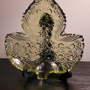 May include: A green glass leaf-shaped dish with a raised, textured pattern. The dish is on a black stand.