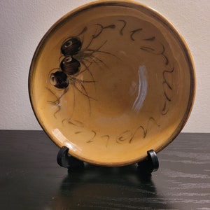 Op de afbeelding: Een ronde, geglazuurde keramische kom met een warme, goudgele afwerking. De binnenzijde is voorzien van een handgeschilderd ontwerp van drie donkerbruine, cirkelvormige figuren met uitstralende lijnen en cursief, donkerbruin schrift langs de rand.