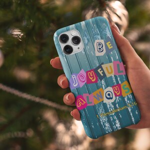 Op de afbeelding: Een blauwe telefoonhoes met een houtnerfpatroon en kleurrijke letters die "Be Joyful Always" spellen met de bijbelverwijzing "1 Thessalonicenzen 5:16".