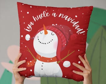 Könnte beinhalten: Rotes Kissen mit einer Schneemann-Illustration und dem Text "¡ya huele a navidad!" in Weiß. Der Schneemann trägt einen roten Hut und einen roten Schal.