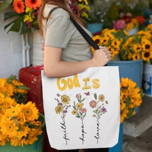 May include: White tote bag with the text "God is" in yellow letters. Below the text are three bouquets of flowers with the words "faith", "hope", and "love" written vertically underneath each bouquet.
