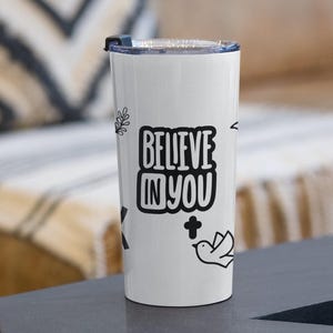 Könnte beinhalten: Ein weißer Edelstahlbecher mit einem schwarz-weißen Design. Der Becher zeigt den Text "Believe in You" mit einer kleinen Vogel- und Kreuzgrafik.