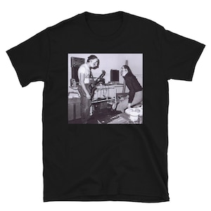 Pode incluir: Fotografia a preto e branco de duas pessoas numa sala. Uma pessoa está a tocar uma guitarra eléctrica, a outra está de pé e a olhar para a pessoa que toca guitarra. A fotografia está impressa numa t-shirt preta.