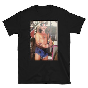 May include: A black t-shirt with a photo of a man wearing a bandana and holding a sword. The man is wearing a blue denim shorts and has a tattoo on his arm.