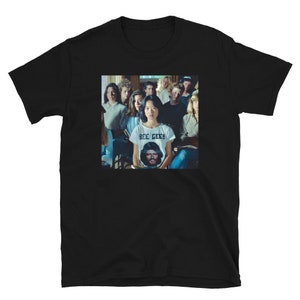 Puede incluir: Camiseta negra con un grupo de personas en un aula. La persona en el centro de la imagen lleva una camiseta blanca con el texto "BEE GEES" impreso.