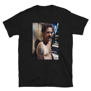 May include: A black and white photo of a man with a mustache and a tattoo on his arm. He is wearing a black shirt and is standing in front of a wooden fence.